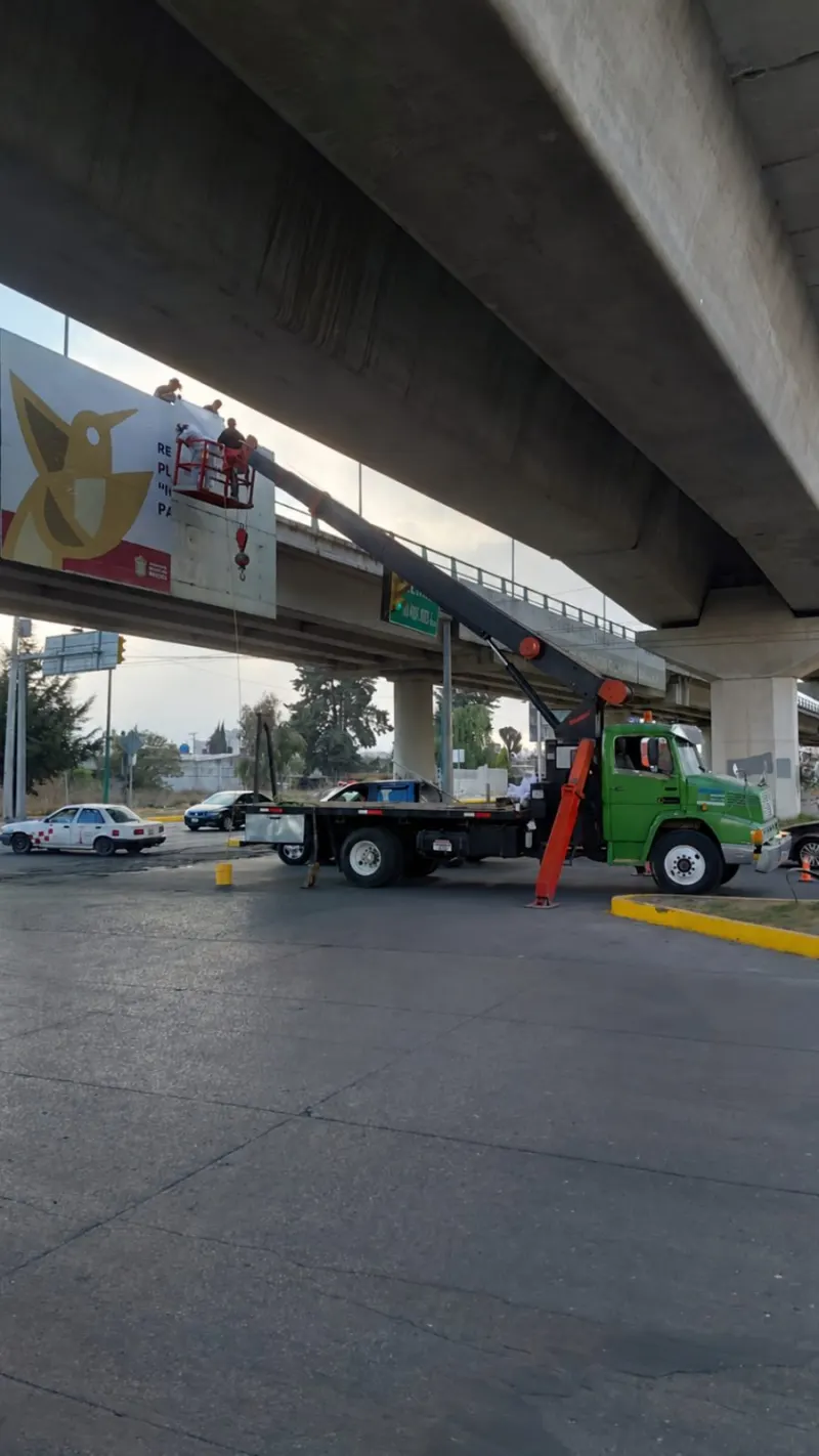Trabajadores en una canastilla roja colocan vinil en un espectacular instalado sobre un puente.