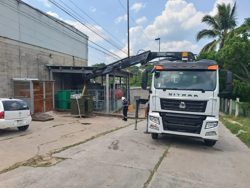 Grúa articulada con un equipo eléctrico suspendido junto a una malla perimetral.