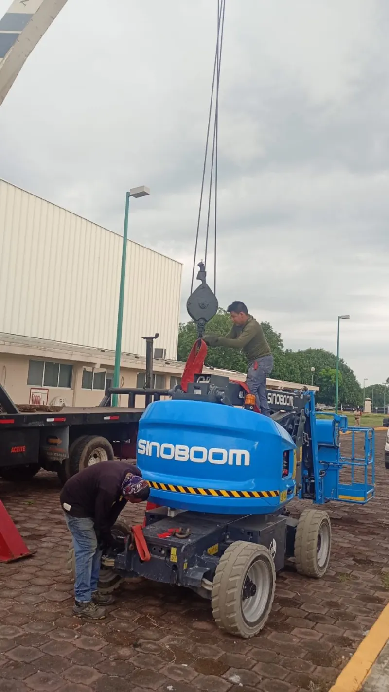 Plataforma elevadora azul sobre piso adoquinado, con dos personas, eslingas y el gancho de la grúa junto a ella.