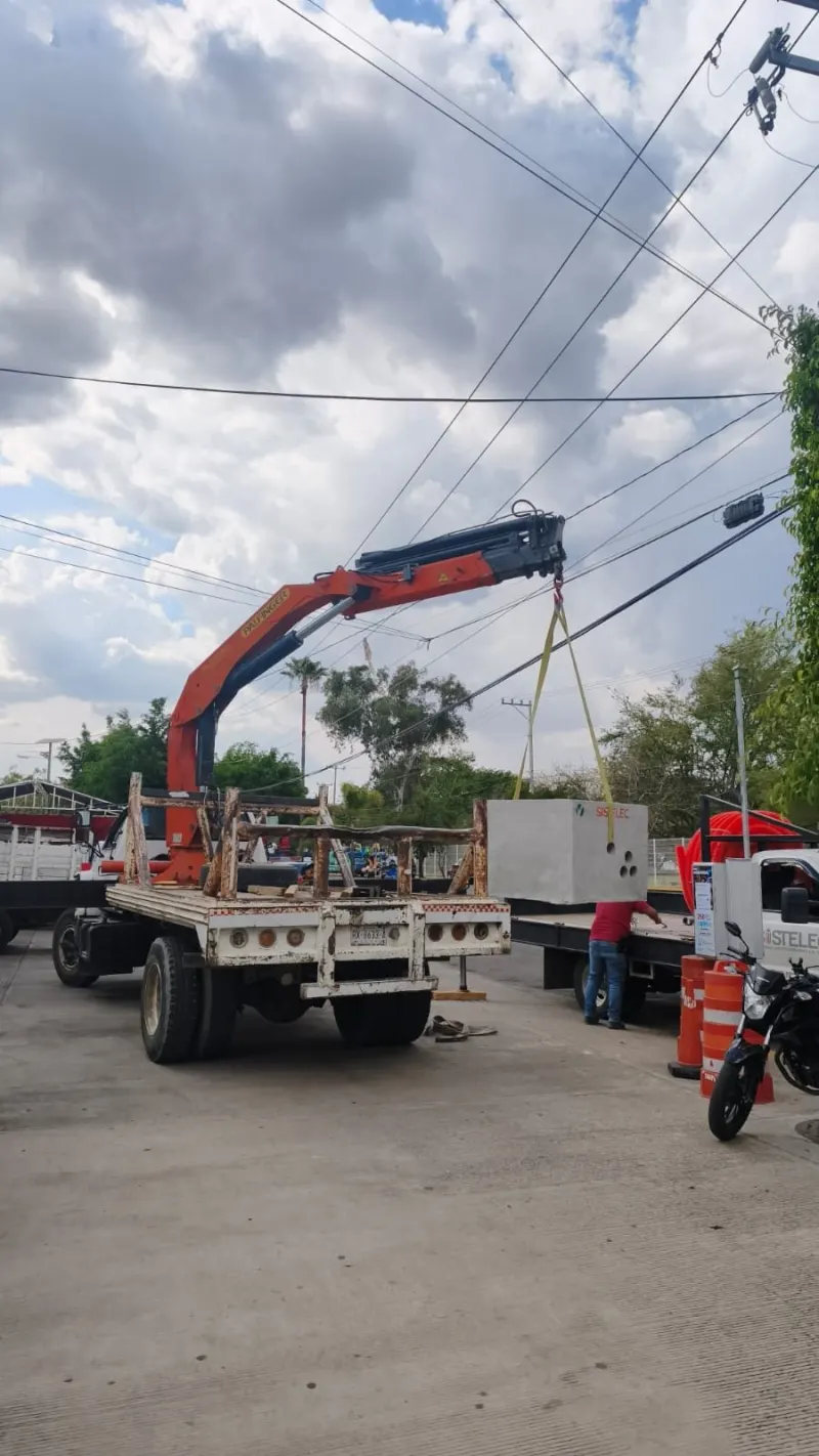 Camión con grúa articulada y pluma elevada entre líneas aéreas junto a una barda metálica.