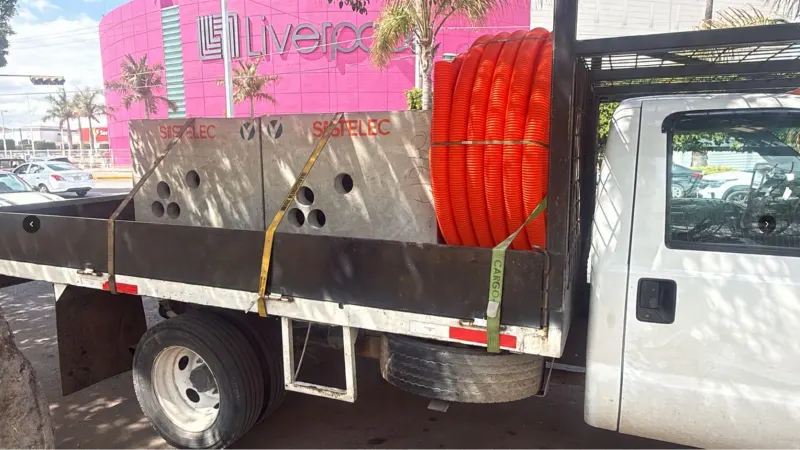Registro de concreto suspendido con eslingas entre dos camiones y bajo líneas aéreas.