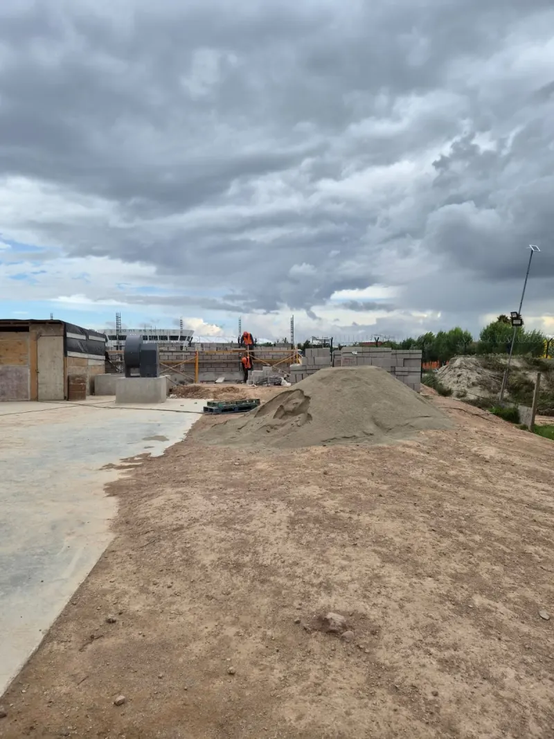 Terreno de tierra con una pila de arena, bloques y un equipo gris sobre una base de concreto.