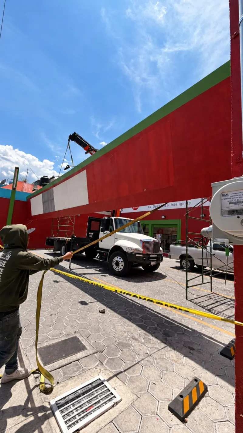 Estructura roja suspendida sobre el camión mientras un trabajador controla una banda amarilla.