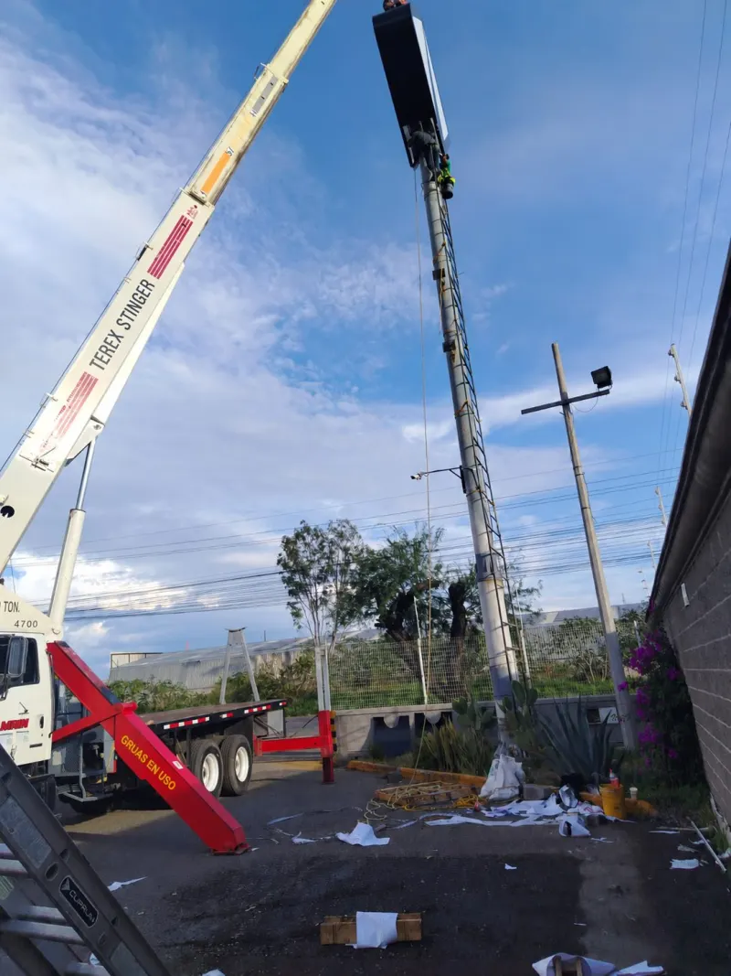 Pluma de grúa junto a una pantalla elevada, con personal en la parte superior de la estructura.