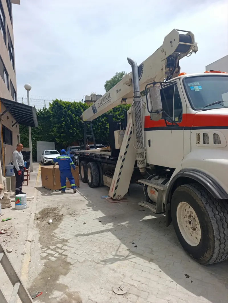Grúa Titán con estabilizador desplegado junto a un aire acondicionado en caja y varias escaleras.