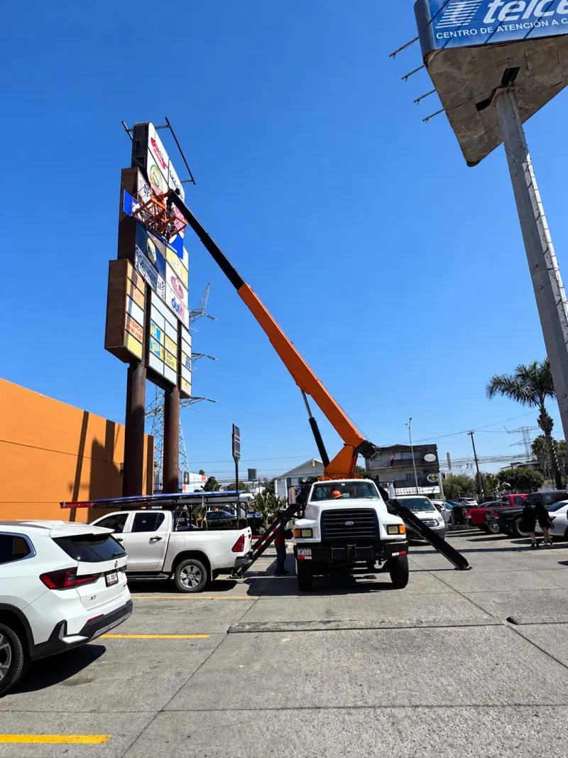Grúa Titán con canastilla elevada junto a un anuncio tipo tótem en un estacionamiento con vehículos.