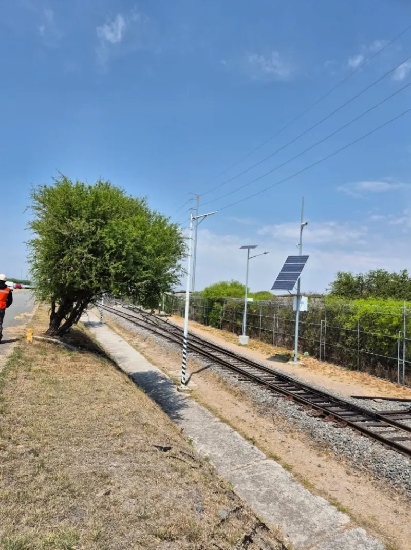 Vías del tren junto a postes con paneles solares y líneas aéreas, vistas desde el camino.