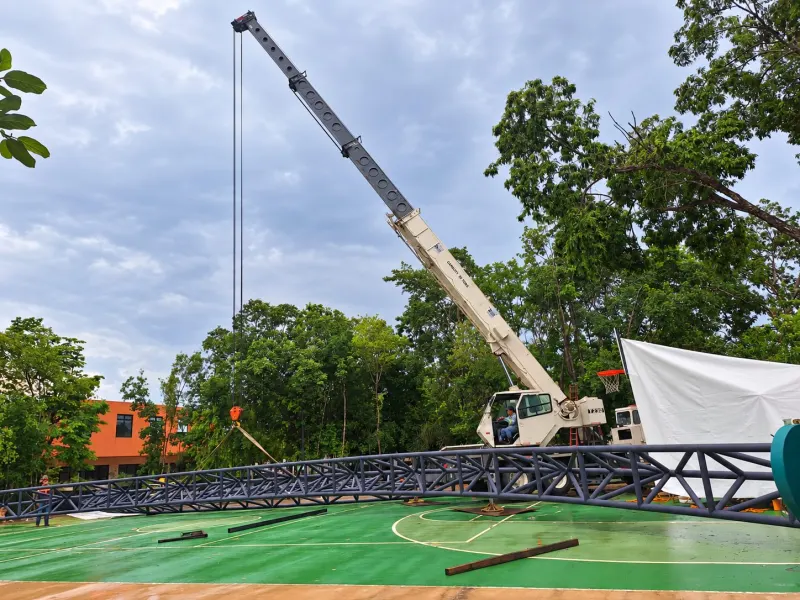 Grúa telescópica junto a una armadura tritubular extendida sobre la cancha verde.
