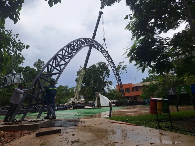 Armadura curva elevada por una grúa mientras personal guía un extremo junto al apoyo sobre piso mojado.