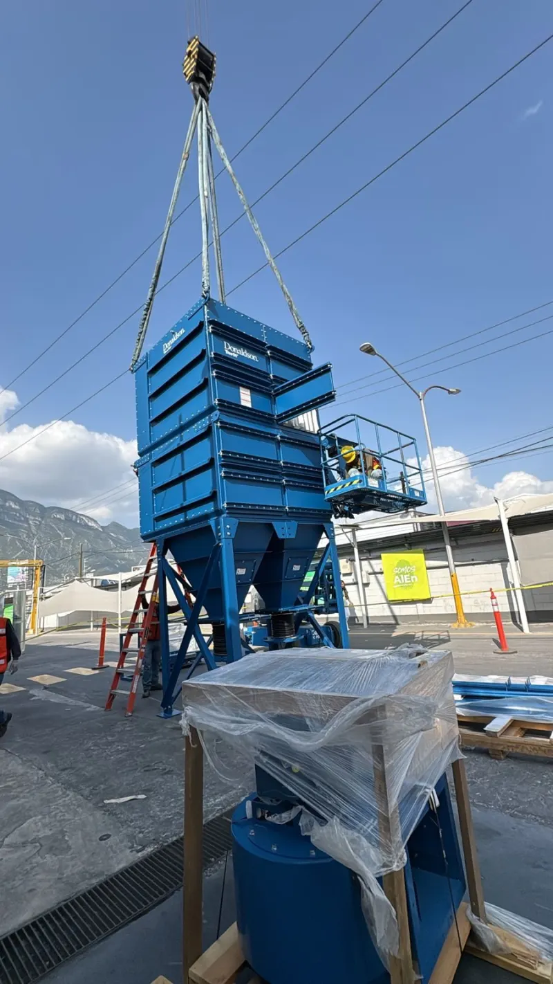 Colector azul suspendido por eslingas junto a una plataforma elevadora en un patio industrial.