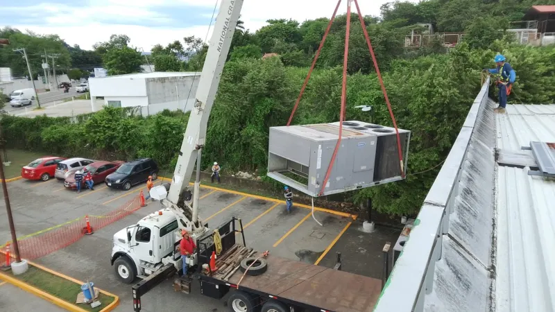 Equipo HVAC gris suspendido con eslingas rojas de una grúa junto al borde de la azotea y cajones de estacionamiento.
