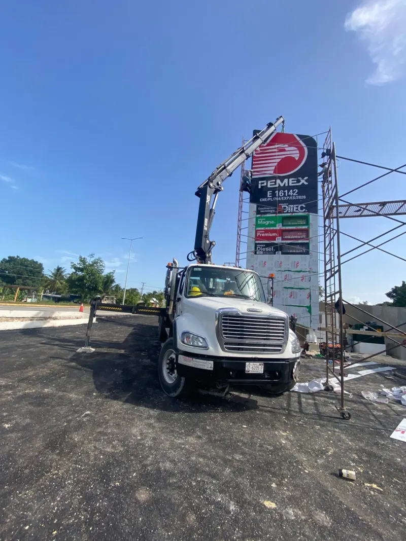 Grúa tipo HIAB con estabilizadores desplegados junto a un tótem rodeado de andamios.