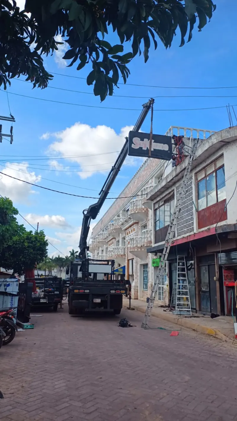 Anuncio negro suspendido por una grúa HIAB frente a una fachada, con cables aéreos y escaleras.