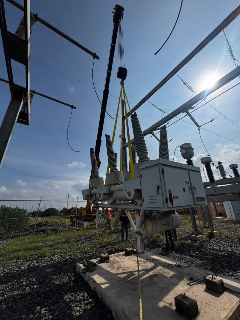 Equipo eléctrico con aisladores negros sujeto por eslingas amarillas frente a una grúa telescópica.