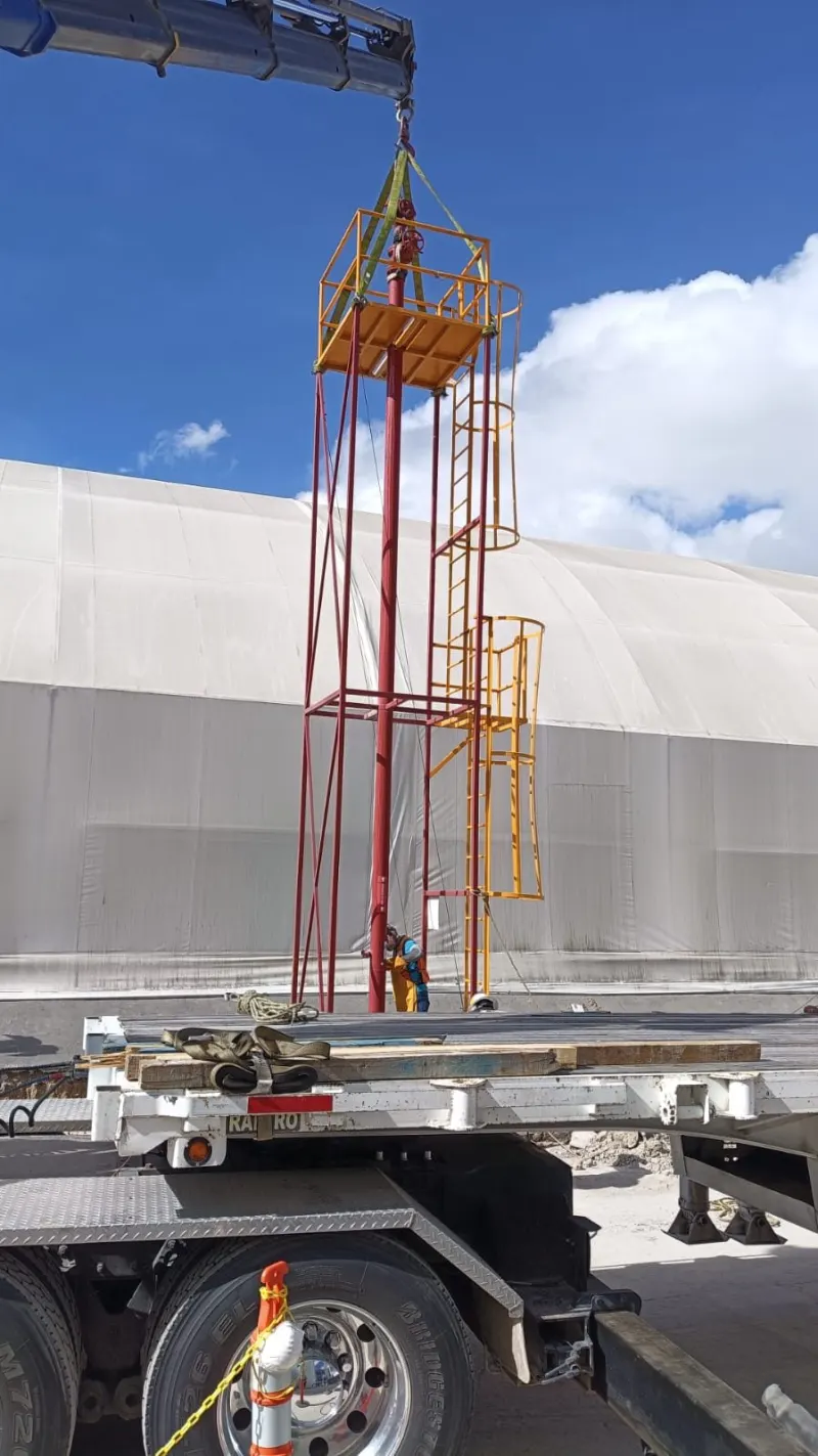 Grúa articulada mantiene vertical una estructura roja con plataformas y escalera junto a un remolque.