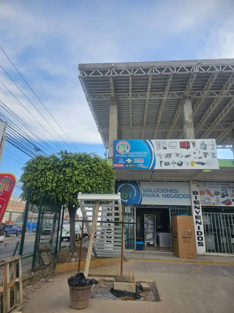 Área comercial con la cimentación del poste frente a un establecimiento y líneas aéreas cercanas.